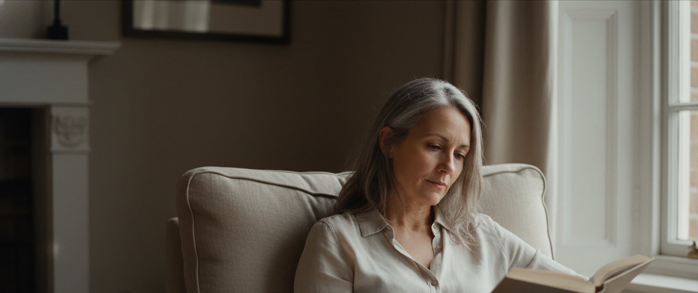 A woman in her early 50s with natural grey hair seated in a cream armchair reading a book, soft daylight from a tall window, a fireplace mantel just visible to one side — quiet, real skin, unposed.